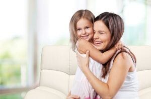 A mom holding her daughter while sitting on a couch. Both are smiling and the mom is laughing.